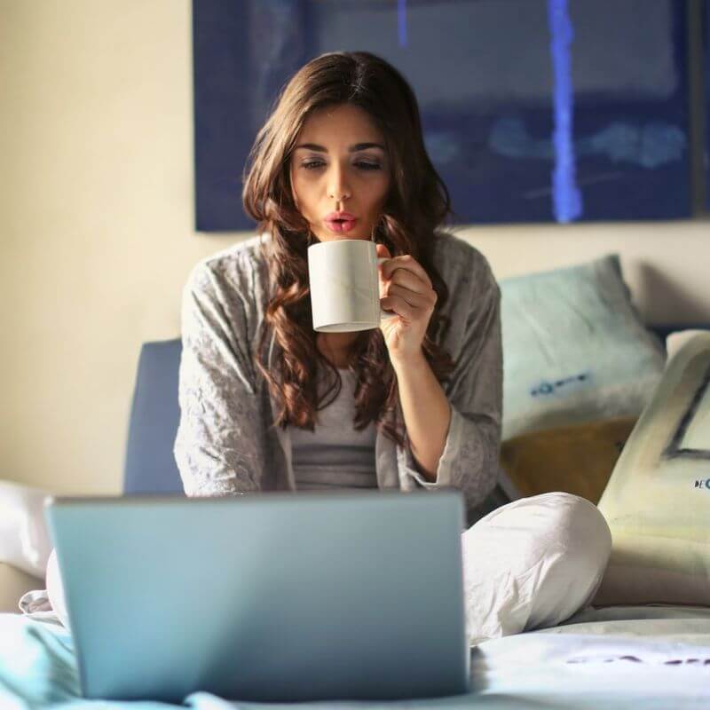 A woman sitting on her bed drinking a hot beverage while working on her laptop