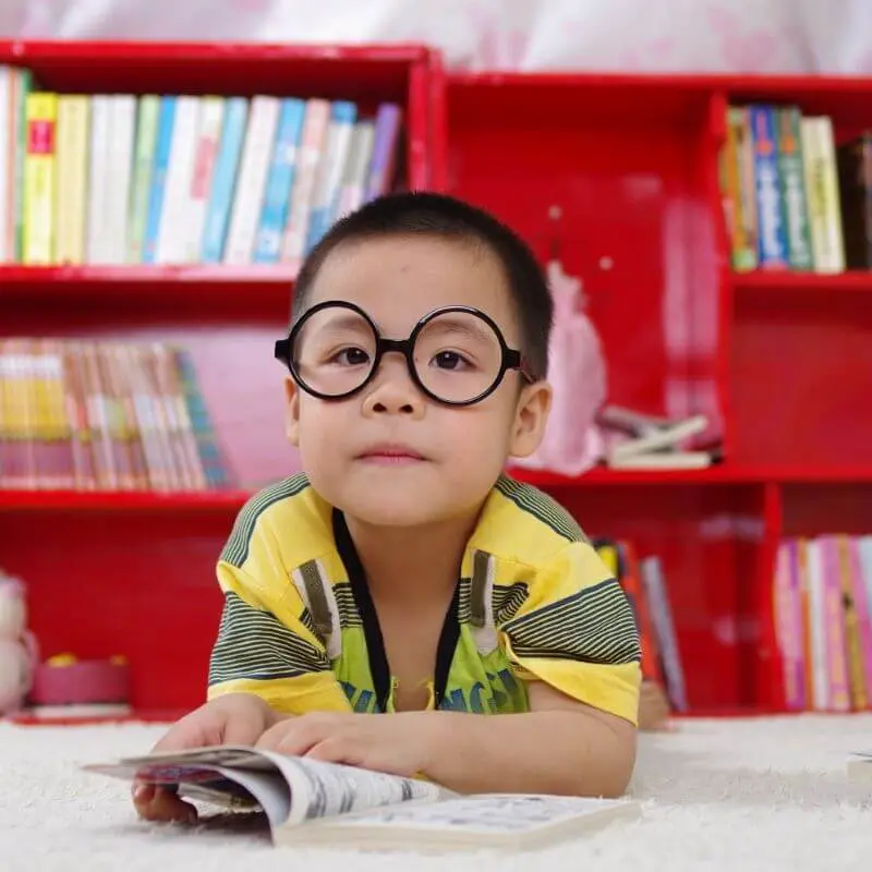 A little boy with glasses in front of a red bookcase