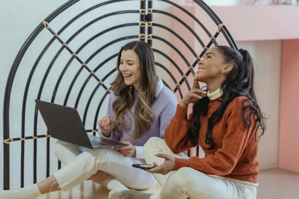 two women sitting together doing remote work on laptop and phone